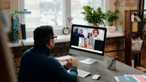 Person in a home office on a video call with four participants on a desktop screen, GoFranchise logo visible on the window sill side.