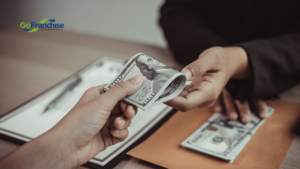 Close-up of hands exchanging cash during a business transaction, GoFranchise logo visible in the top-left corner.