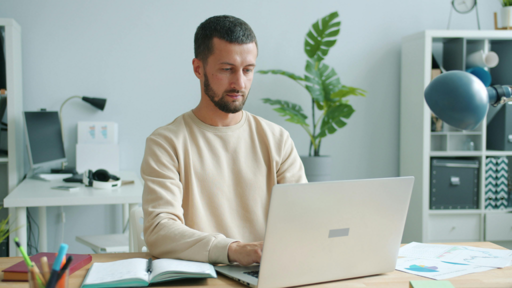 A man sits at a desk working on a laptop, with an open notebook and scattered documents nearby in a bright home office.