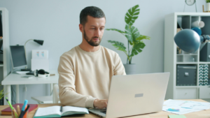A man sits at a desk working on a laptop, with an open notebook and scattered documents nearby in a bright home office.